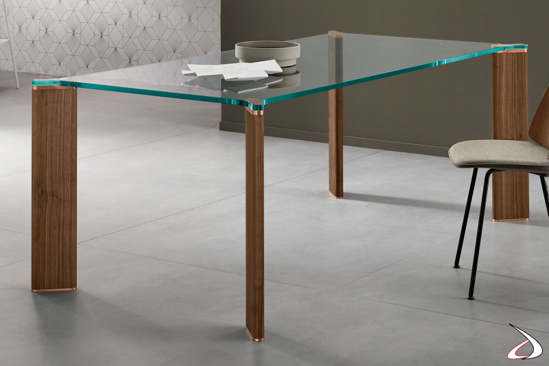 Tavolo moderno e di design caratterizzato dal top in vetro e dalle gambe in noce canaletto con piastre terminali in rame.